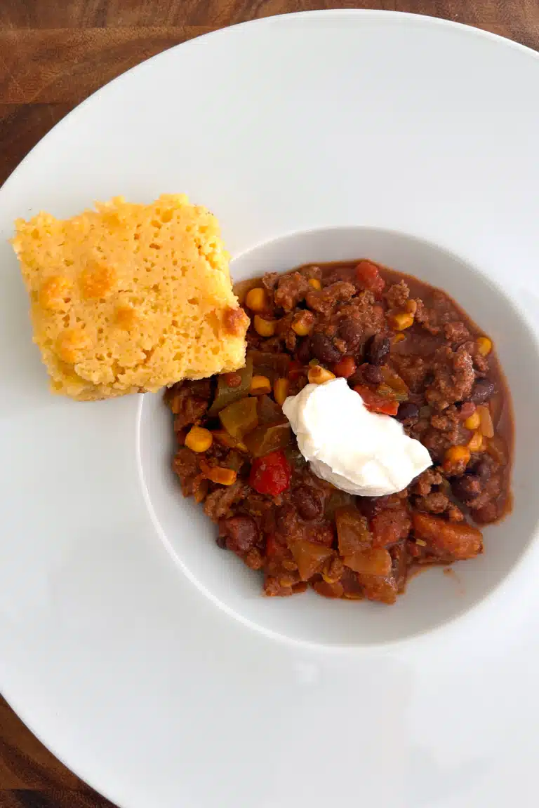 The best beef chili in a white bowl topped with sour cream and a side of cornbread.